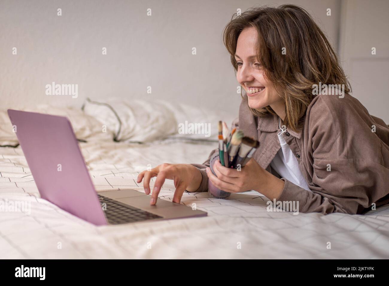 Photo d'une fille souriante enthousiaste se préparant à apprendre à peindre avec des leçons en ligne via son ordinateur portable avec un pot de pinceau de sa salle Banque D'Images