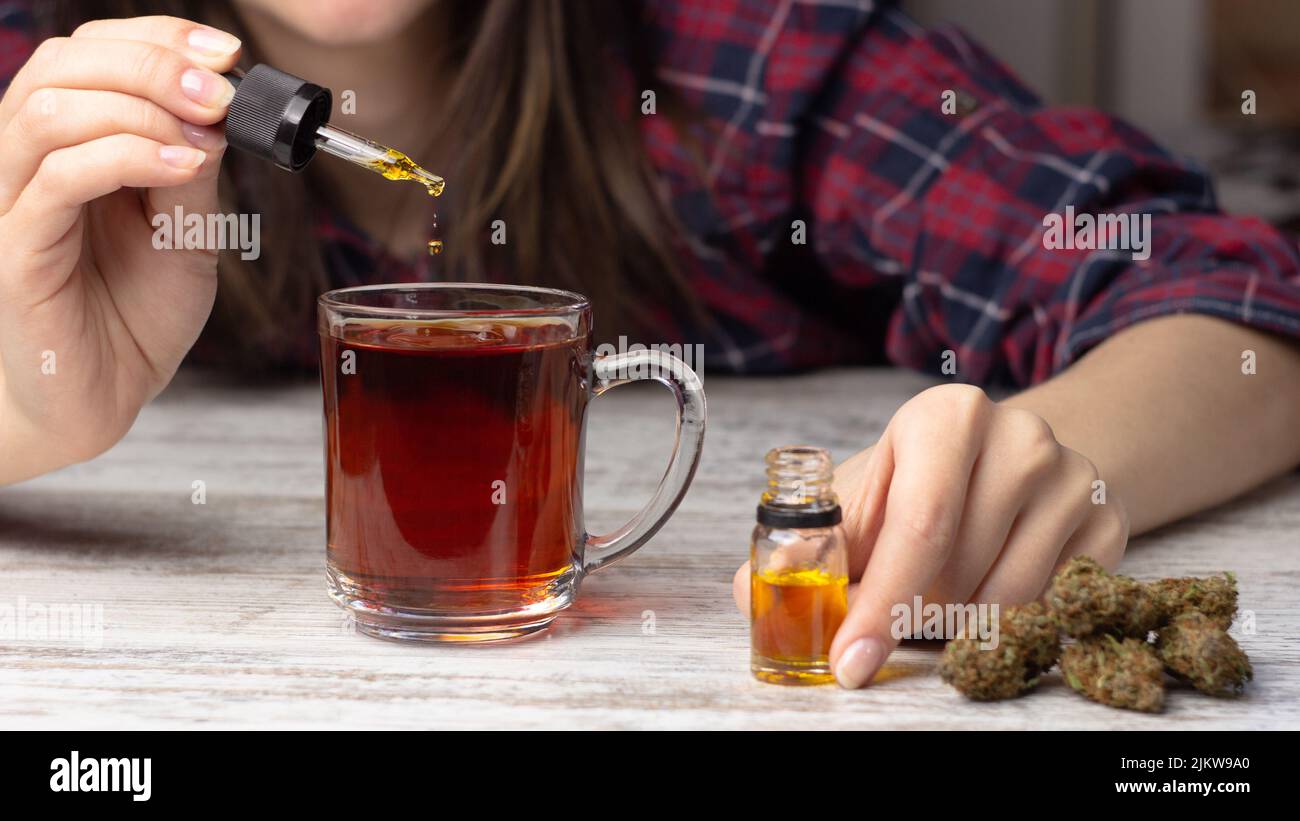Les mains des femmes ajoutent de l'huile de marijuana cbd d'une pipette au thé. Plusieurs bourgeons secs de cannabis médical se trouvent à proximité Banque D'Images