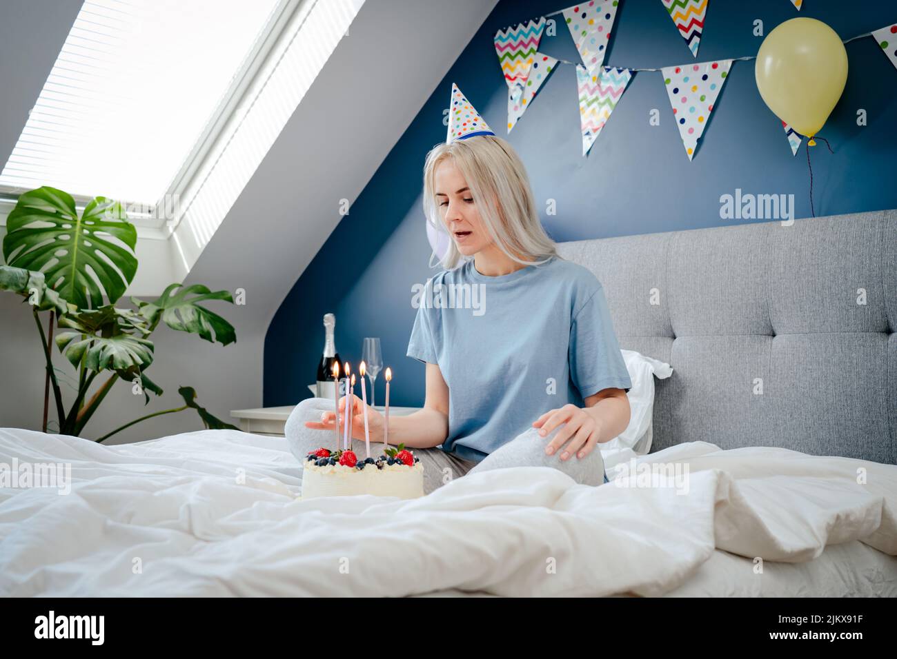 Femme attentive en chapeau de fête d'anniversaire avec gâteau de fête avec bougies sur le lit dans la chambre décorée. Faites un souhait. Surprise du matin. Le bi heureux Banque D'Images Femme attentive en chapeau de fête d'anniversaire avec gâteau de fête avec bougies sur le lit dans la chambre décorée. Faites un souhait. Surprise du matin. Le bi heureux Banque D'Images