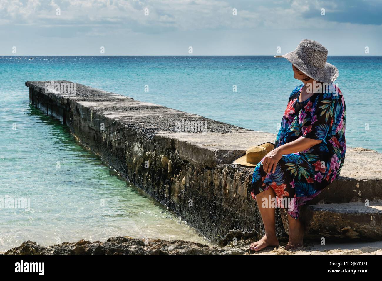 Femme d'âge moyen assise sur un petit quai en pierre dans une plage des Caraïbes avec des eaux calmes et une mer bleu clair, il ya un ciel nuageux, elle regarde loin Banque D'Images