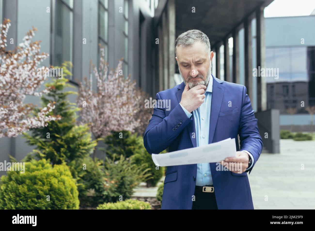 Homme de chef senior et expérimenté à cheveux gris près du bureau pensif lisant le document de rapport financier à l'extérieur du bureau, homme d'affaires pensant Banque D'Images