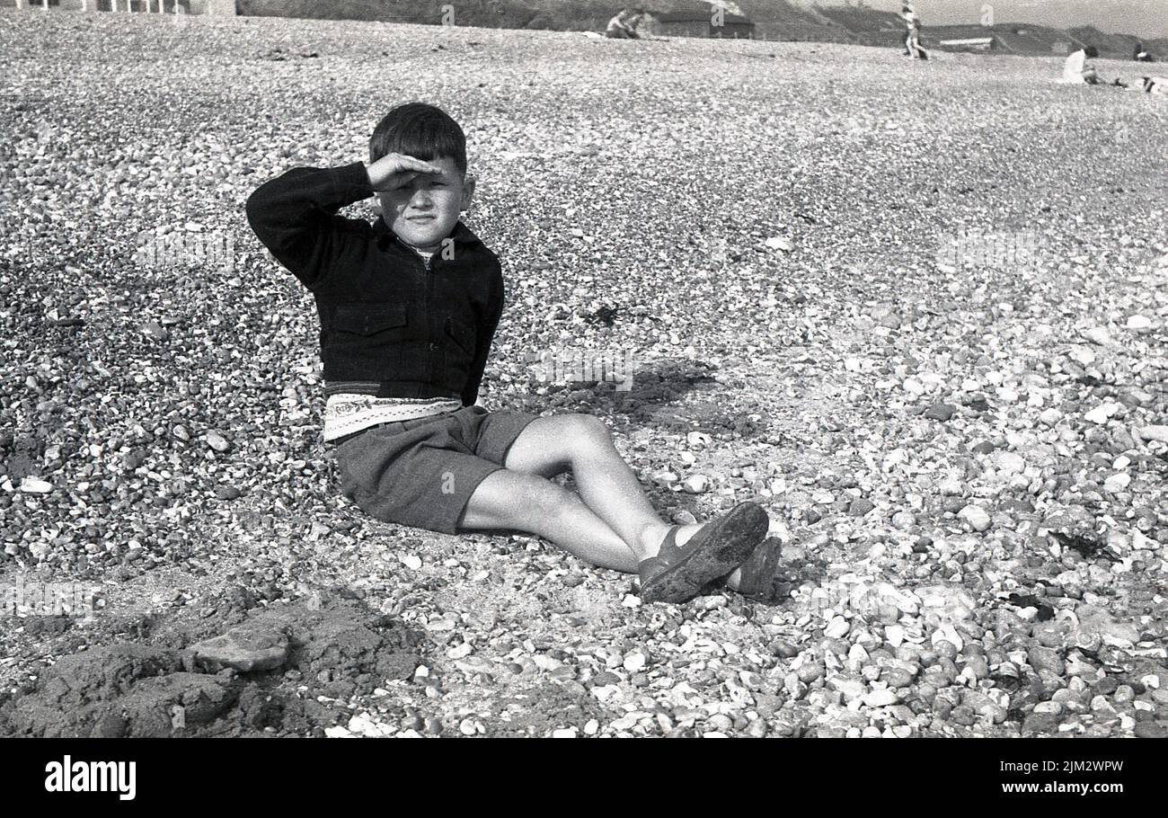 1960s, historique, un garçon assis sur une plage de galets, avec vue sur la mer, Angleterre, Royaume-Uni. Banque D'Images