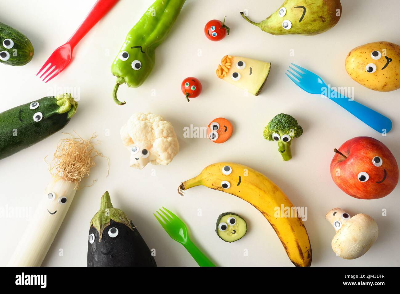 Arrière-plan avec une variété de fruits et légumes drôles présentés pour la nutrition des enfants avec les yeux et la bouche sur une table blanche avec des fourchettes colorées. Banque D'Images