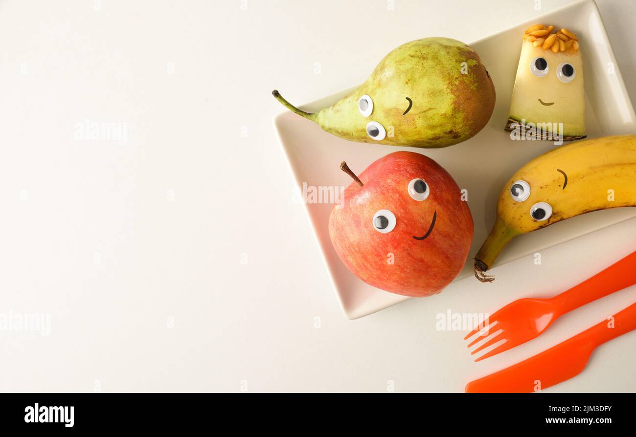 Fond avec assiette de fruits drôles avec yeux et bouches dessinés servi pour les enfants sur table blanche. Vue de dessus. Banque D'Images
