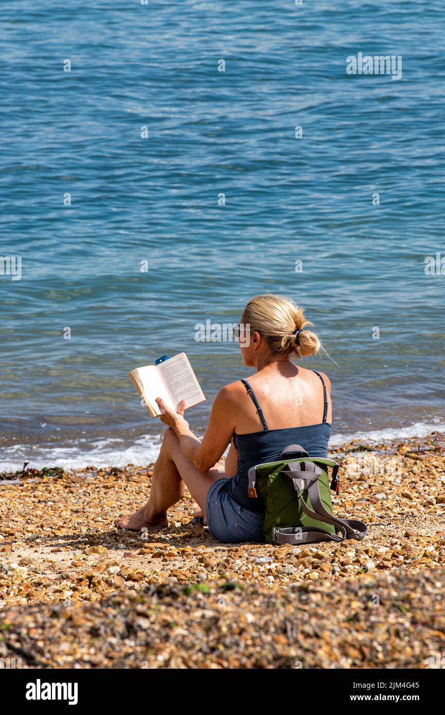 jeune femme attrayante portant une robe d'été lisant un livre assis sur une plage de galets pendant une vague de chaleur d'été, femme assise au soleil sur la plage. Banque D'Images
