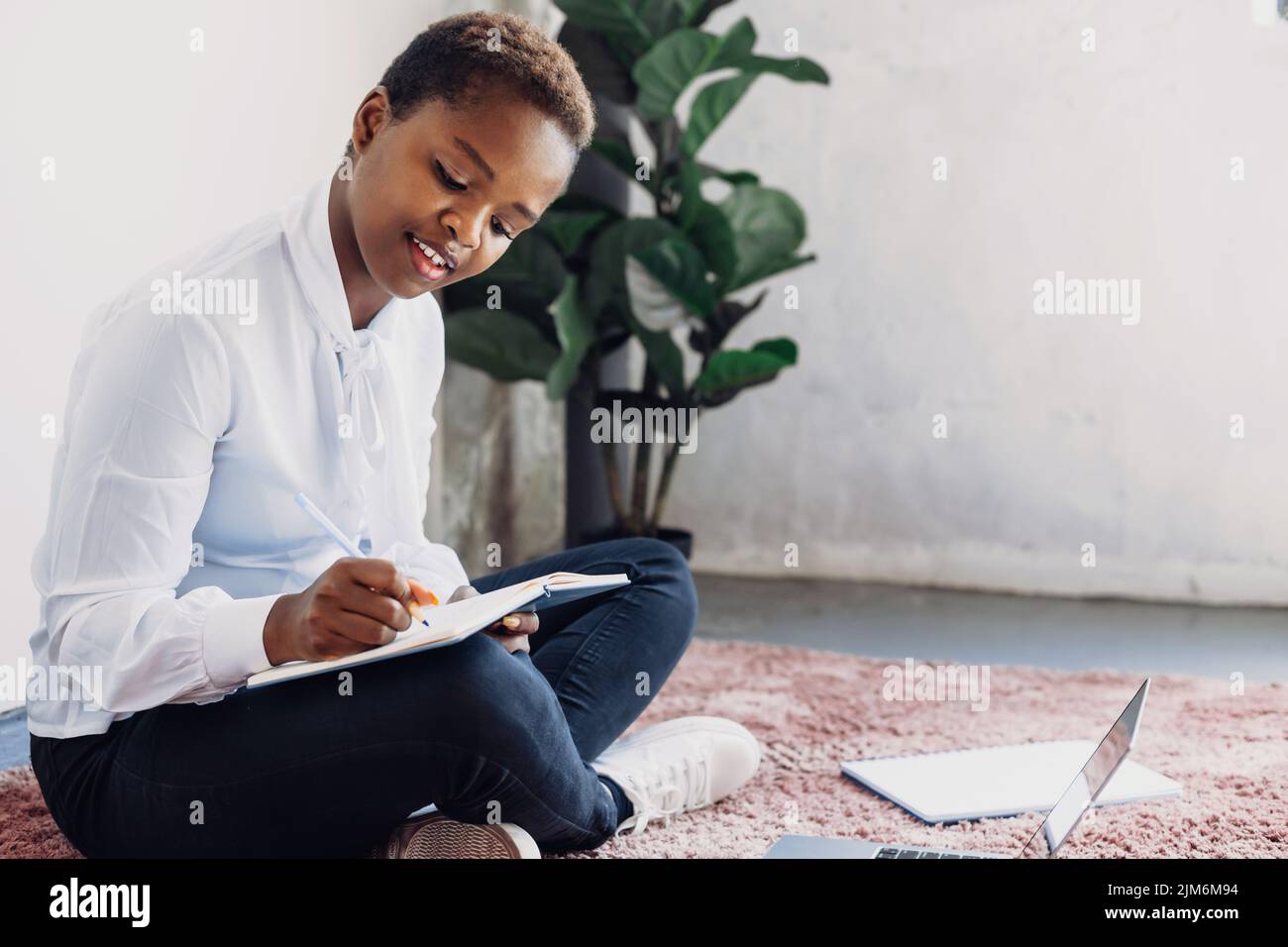 Une jeune fille étudiante afro assise sur le sol préparant ses devoirs pour la leçon de demain à l'université. Cours, réserver, parler sur appel vidéo sur ordinateur portable, écrire Banque D'Images