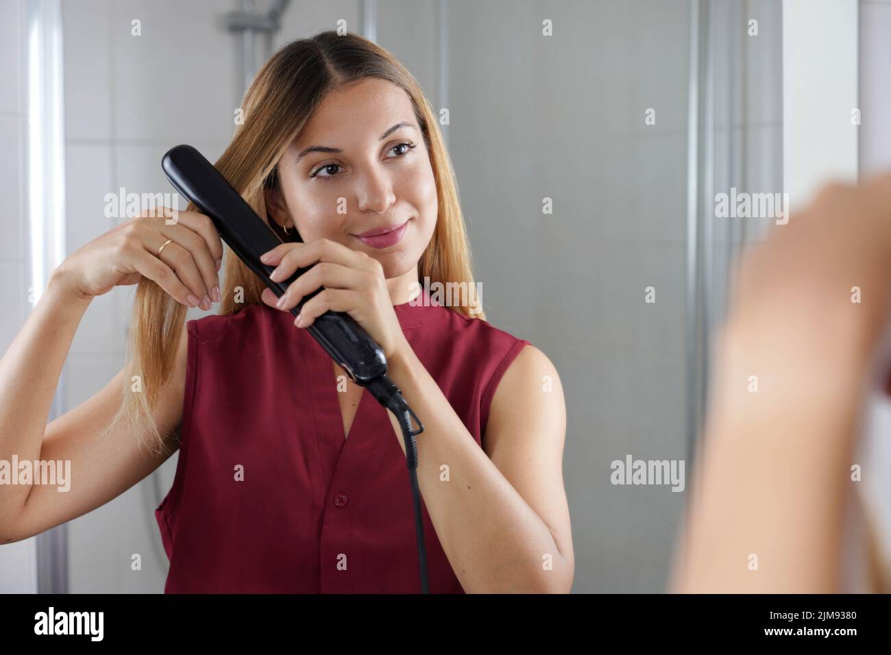 Belle jeune femme utilisant un lisseur vapeur pour coiffer les cheveux au miroir de la salle de bains Banque D'Images