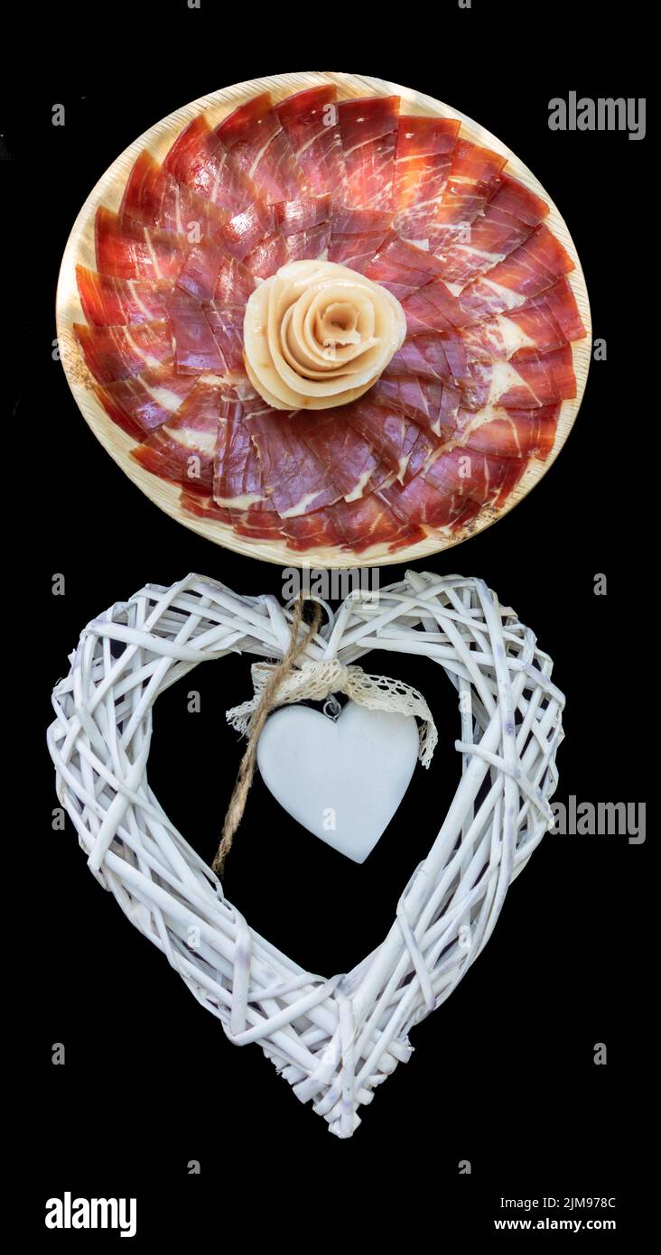 Plato de jamón ibérico recién cortado y en el centro elaborada una rosa con tocino del jamón. Abajo, dos corazones blancos y una puntilla. Banque D'Images