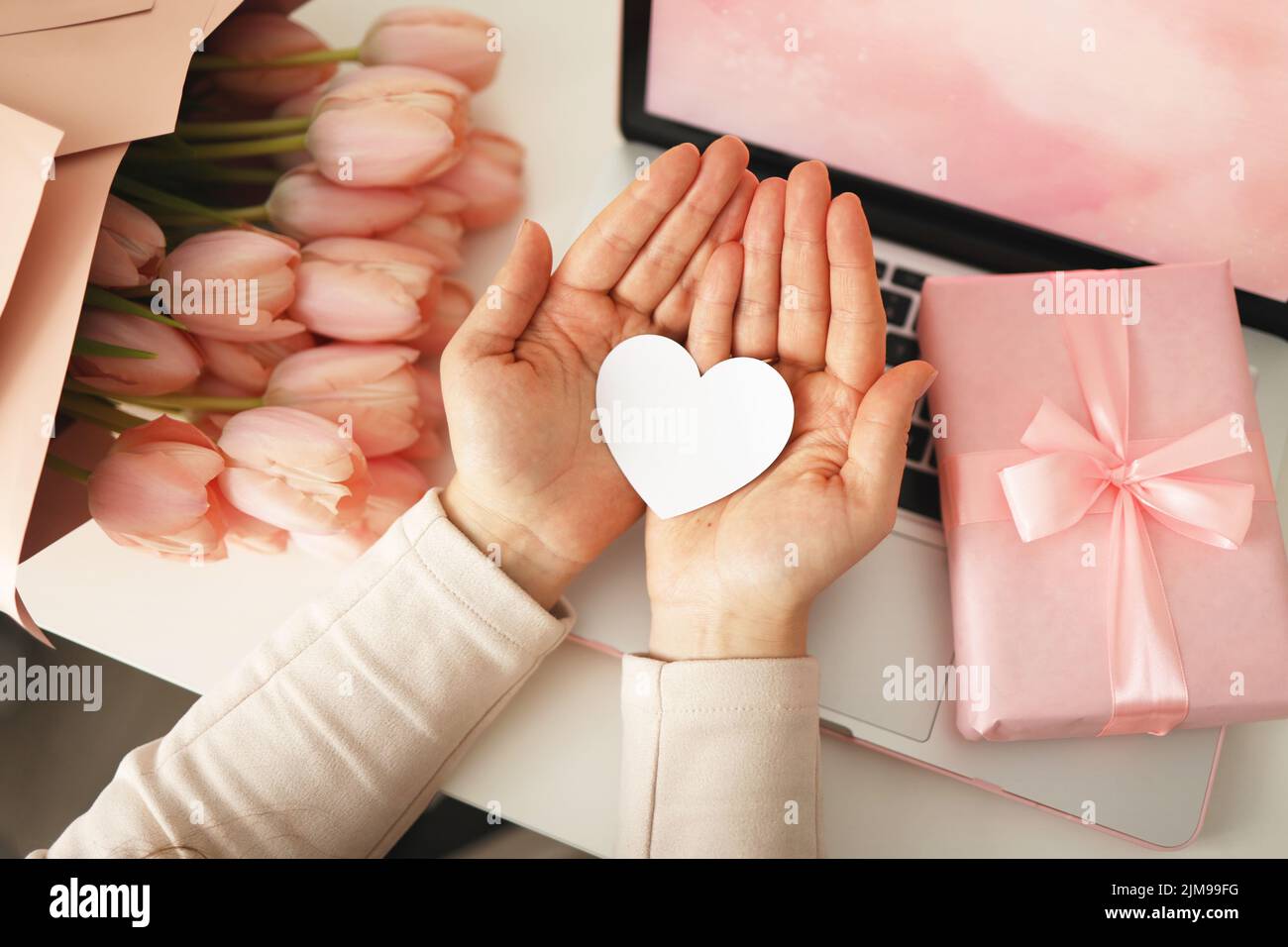 Mains de femme tenant une carte coeur en papier. Fond rose, concept de la Saint-Valentin. Tulipes fleur et boîte-cadeau rose en arrière-plan. Bureau pour femmes. Banque D'Images