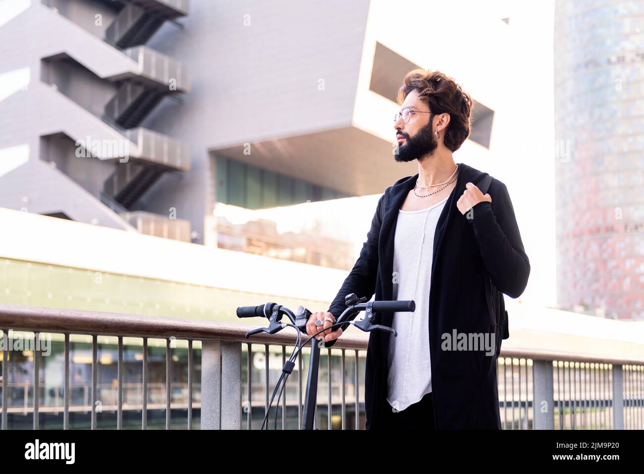 jeune homme caucasien avec barbe portant des vêtements tendance et marchant avec son vélo pliant à travers la ville, concept de style de vie urbain et durable m. Banque D'Images