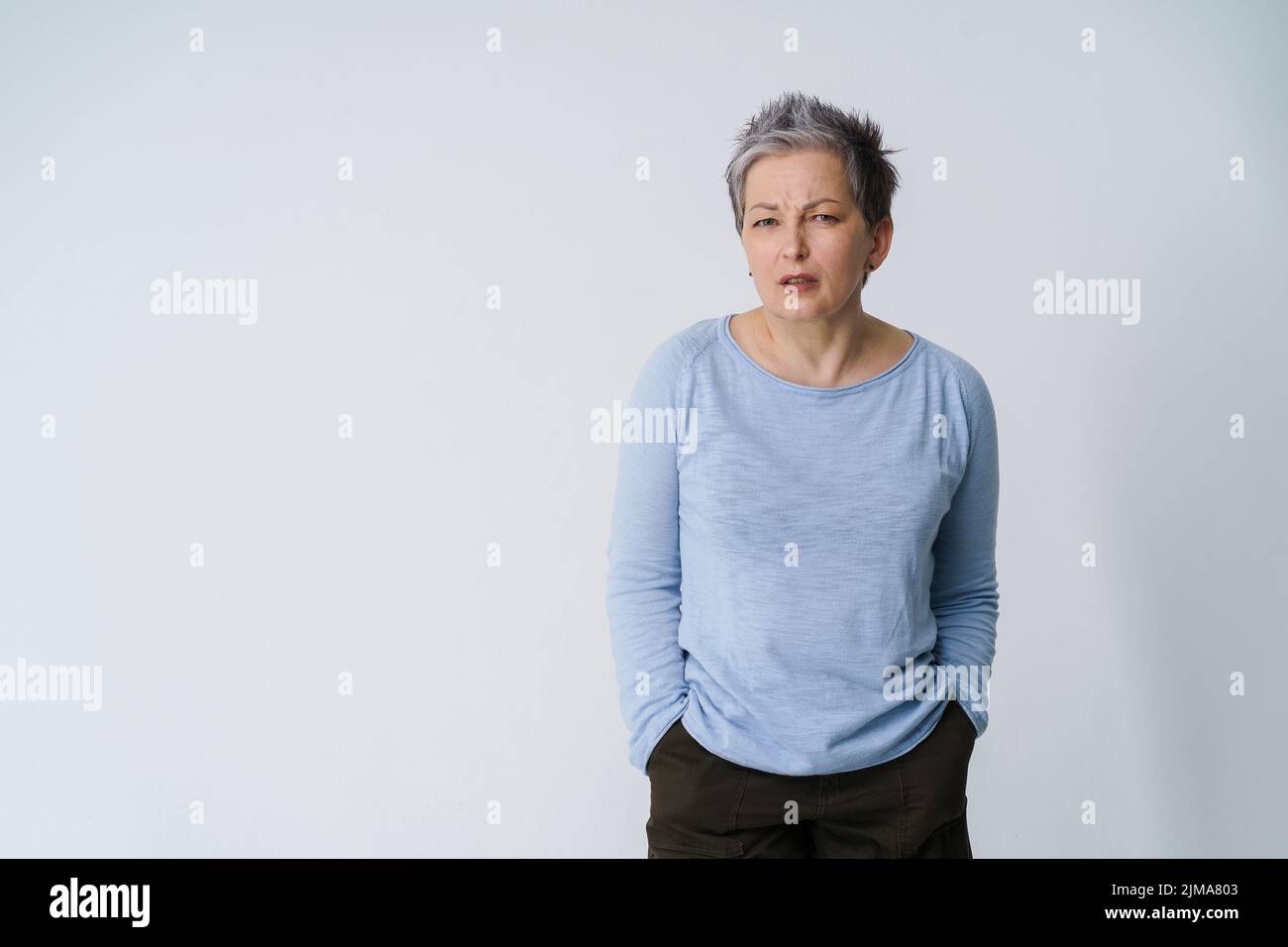 Stressée, en colère mature gris cheveux femme 50s posant frustré regarder la caméra avec les mains dans les poches, copier l'espace sur la gauche isolé sur fond blanc. Soins de santé des personnes matures. Banque D'Images