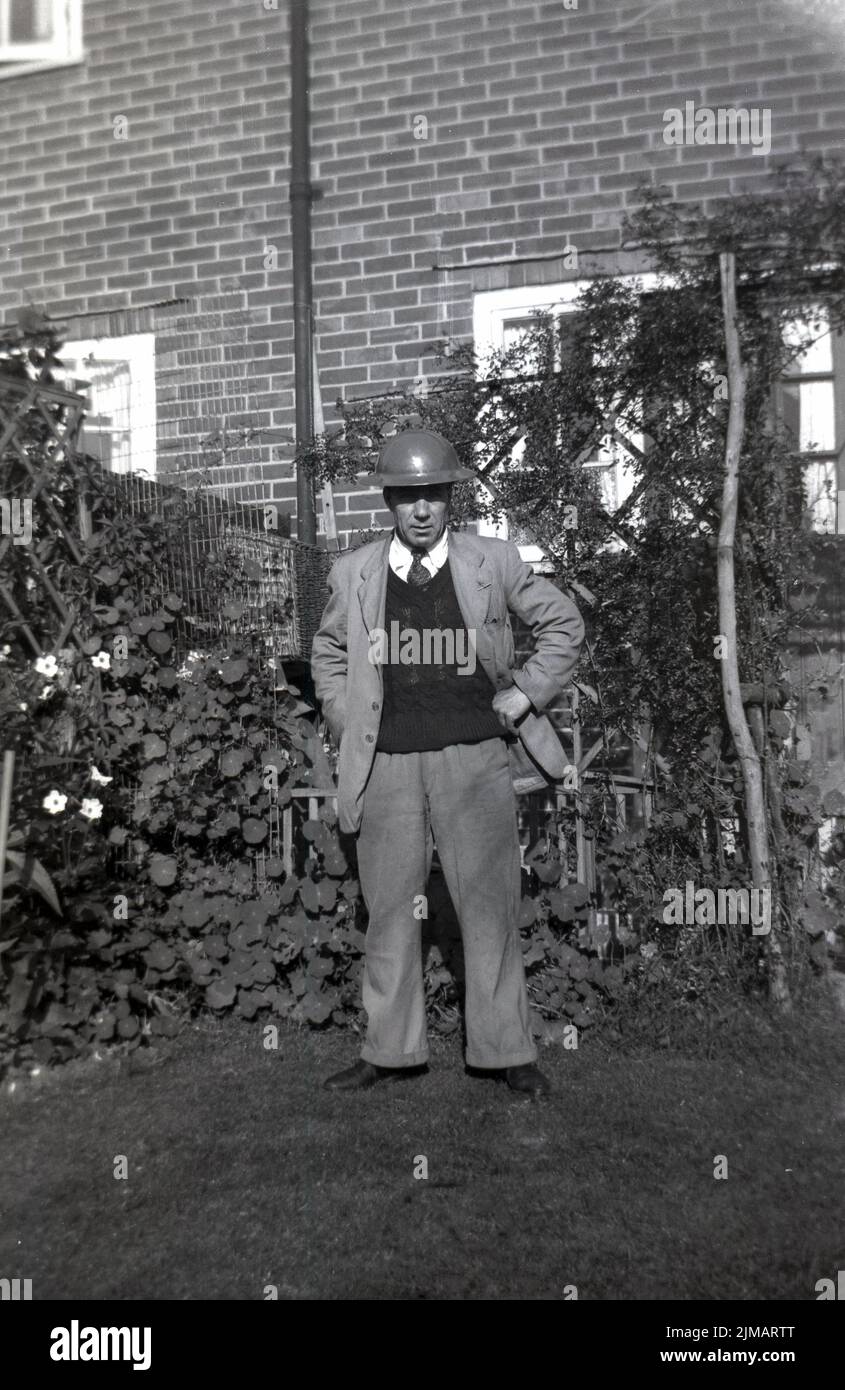 1950s, un homme plus âgé debout dans un jardin à l'arrière, une veste, une cravate et un pull, avec un casque de combat en métal sur sa tête, Angleterre, Royaume-Uni. Un casque de combat en acier, connu sous le nom de 'casque Brodie', d'après son créateur John Leopold Brodie, qui a breveté son design original en 1915, des versions modifiées ont été connues sous le nom de : Battle Bowler, tommy Hat et TiN Hat. Les chapeaux de combat en métal ont été créés peu après le début de WW1, car le casque en tissu et en cuir que les solives portaient traditionnellement n'offrait aucune protection contre l'artillerie. Le casque a également été porté en WW2, ainsi que par la police, les pompiers et les gardes du FSIA. Banque D'Images