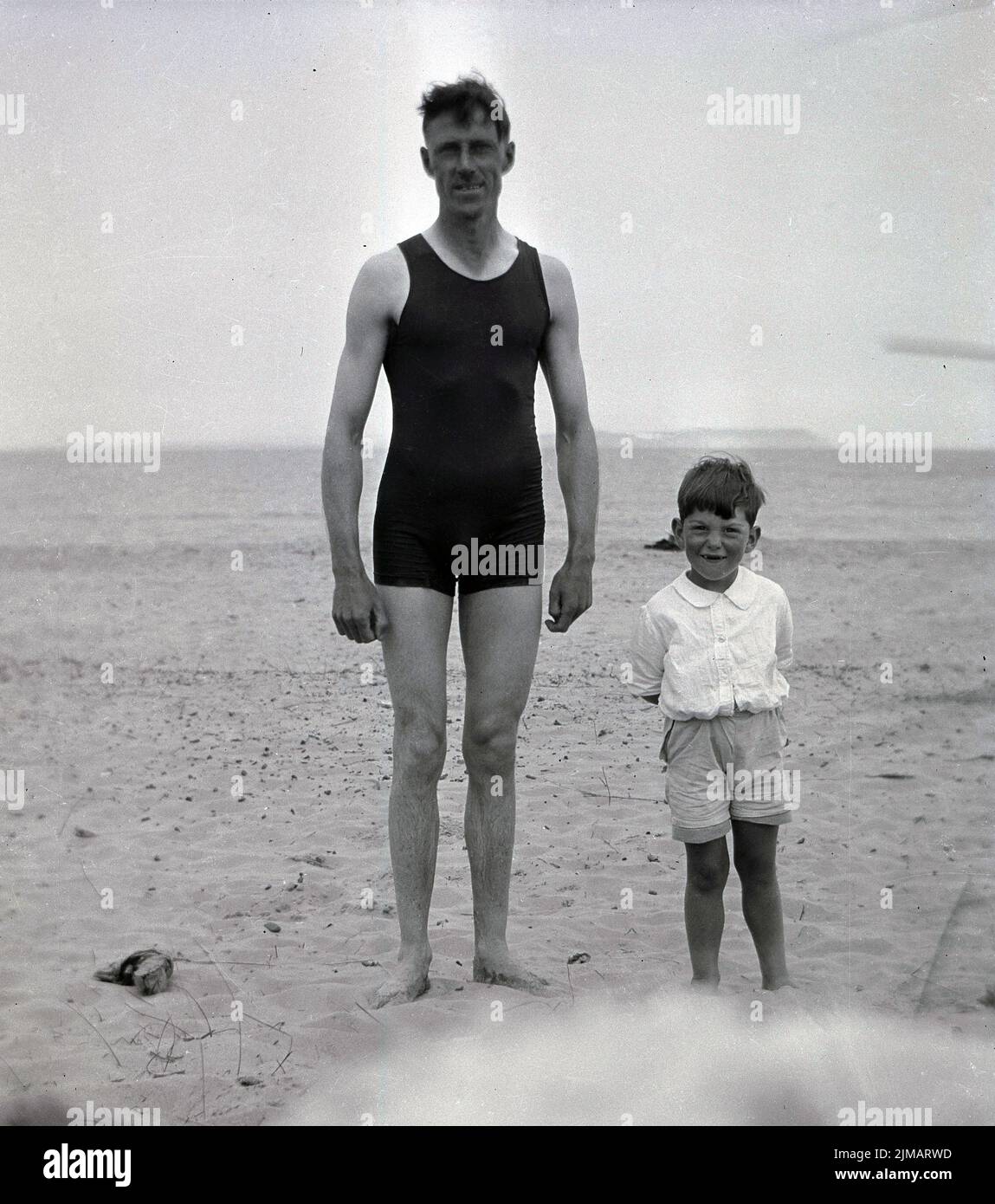 1930s, historique, un père, dans un costume de natation sans manches d'une pièce de l'époque, debout pour une photo sur une large plage de sable avec son petit fils, Angleterre, Royaume-Uni. Banque D'Images