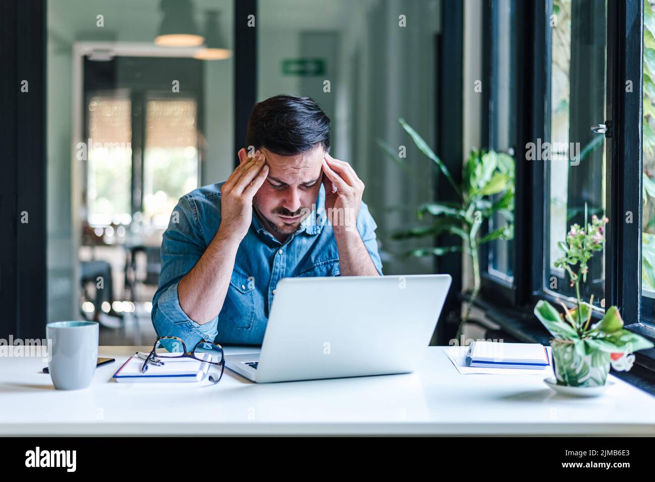 Entrepreneur stressé travaillant sur ordinateur portable. Jeune professionnel d'affaires assis avec la tête dans les mains. Il porte des vêtements décontractés tout en travaillant près de la fenêtre Banque D'Images