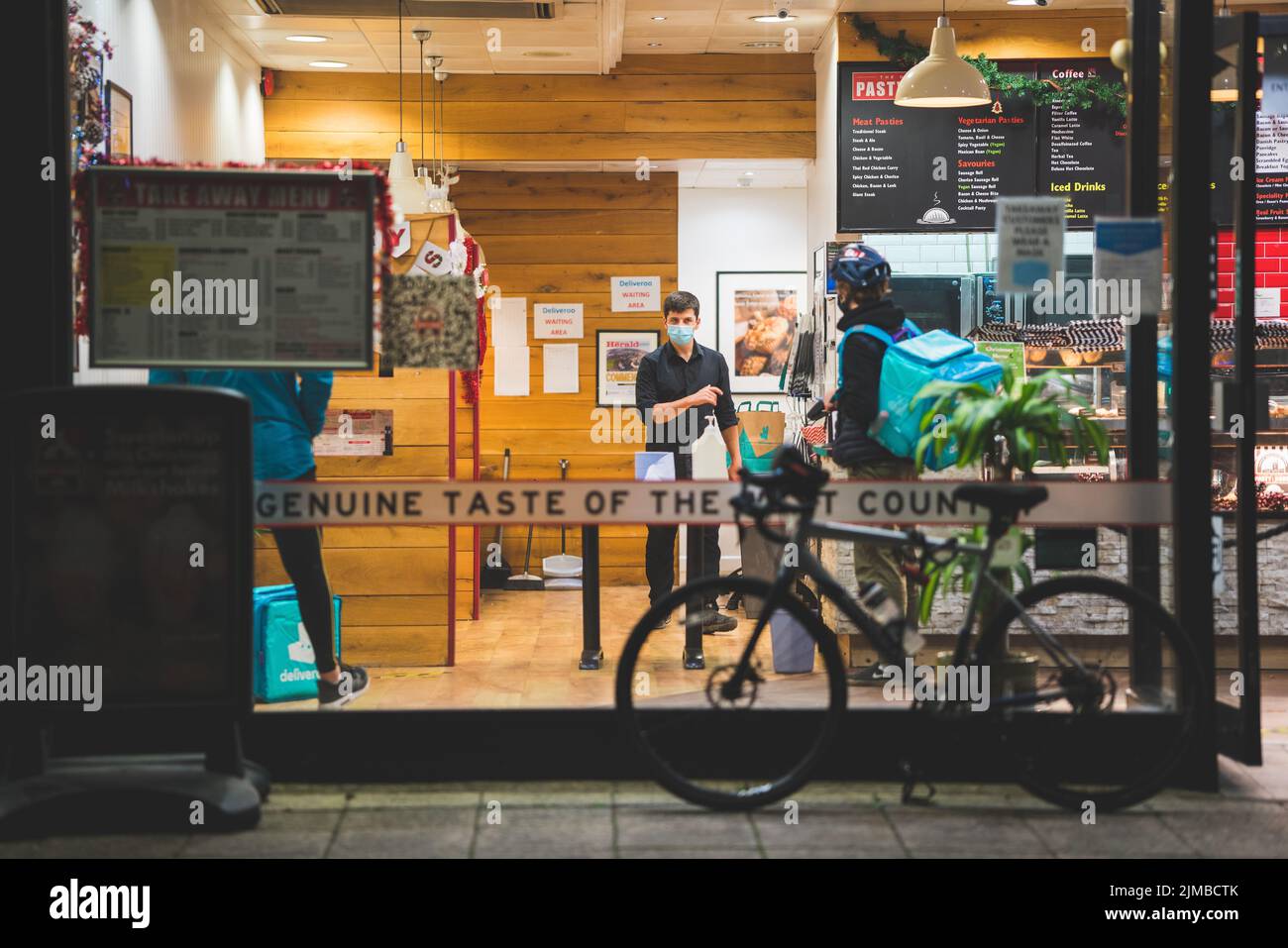 Un pilote de livraison Deliveroo collecte une livraison de nourriture d'un magasin local de restauration rapide Pasty un autre pilote attend la nuit Banque D'Images