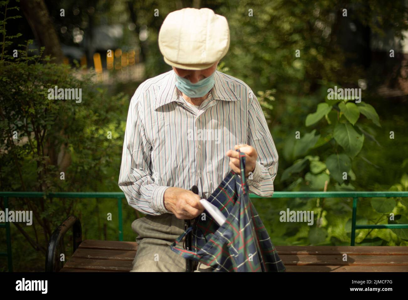 Le vieil homme est assis sur le banc en été. Retraité en Russie près de maison. Homme portant un masque médical. Grand-père en chemise et casquette. Banque D'Images