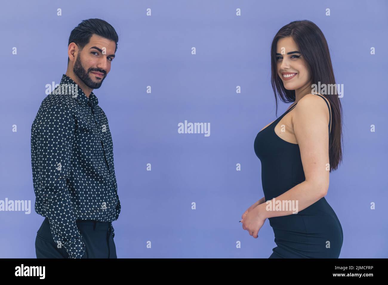 Homme hispanique portant une chemise à motifs regardant séductivement dans l'appareil photo debout devant une femme avec de longs cheveux portant robe noire souriante. Prise de vue en studio. Photo de haute qualité Banque D'Images