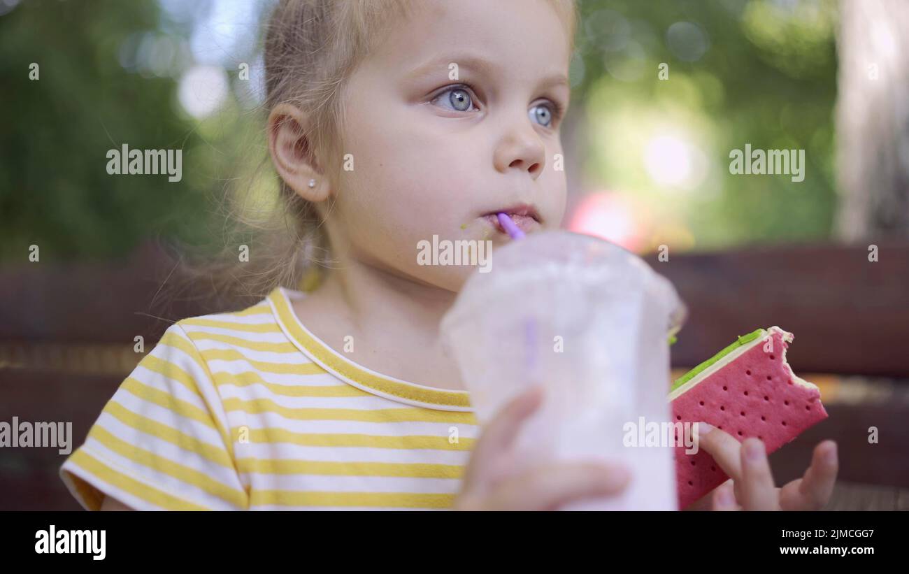 Une petite fille boit un milkshake à travers une paille et saumone à la glace de son autre main. Portrait en gros plan d'une petite fille adorable assise sur un banc de parc Banque D'Images