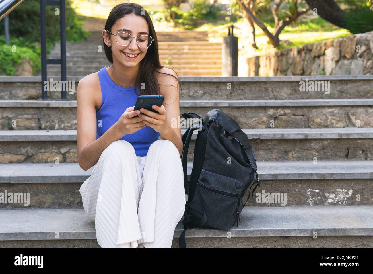 Une étudiante positive envoie un message par SMS au téléphone portable tout en étant assise dans un escalier avec sac à dos dans la rue de la ville le jour de l'été Banque D'Images