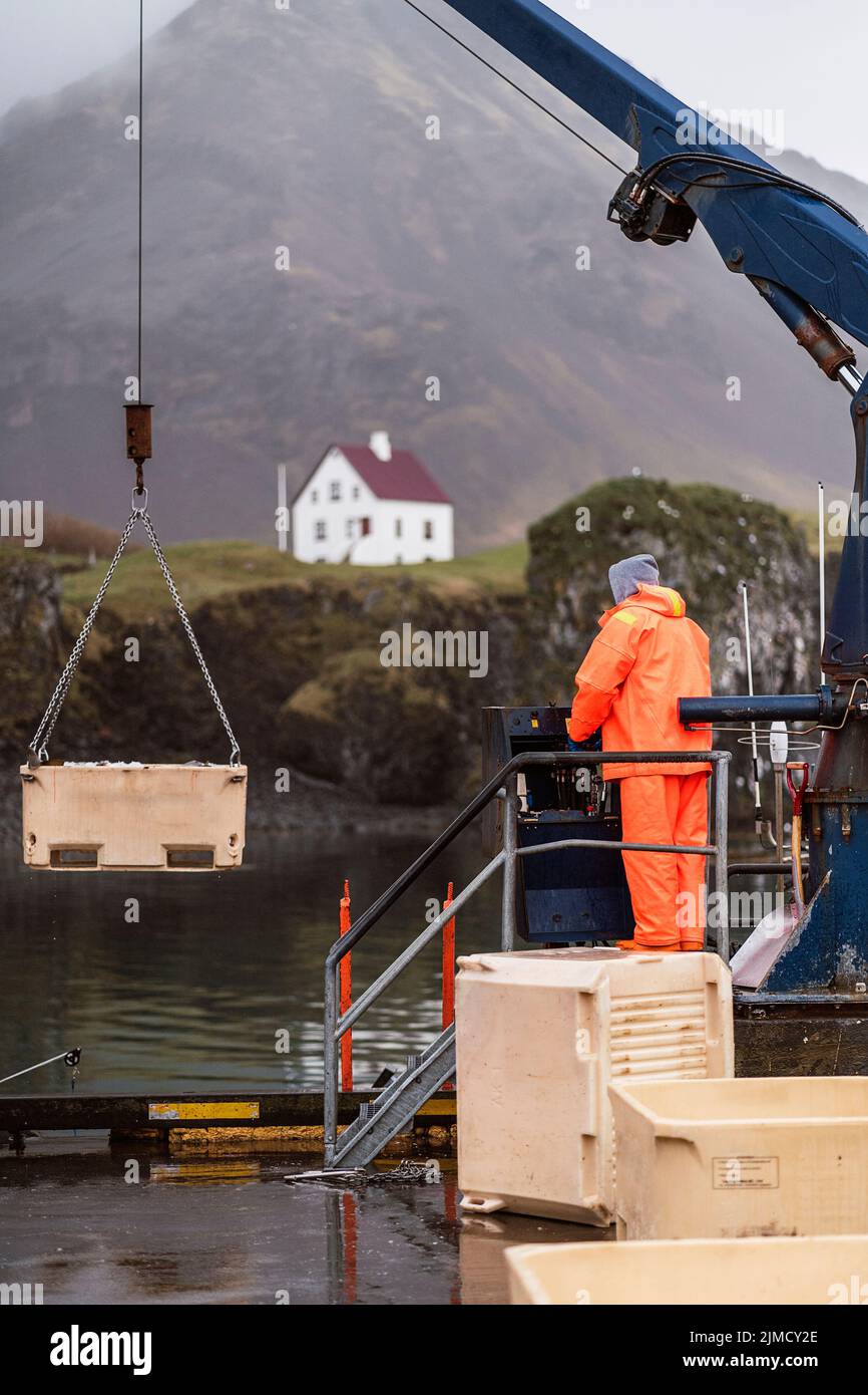 Homme anonyme en uniforme orange commandant la grue pendant le travail sur la ferme piscicole le jour gris dans les montagnes de l'Islande Banque D'Images