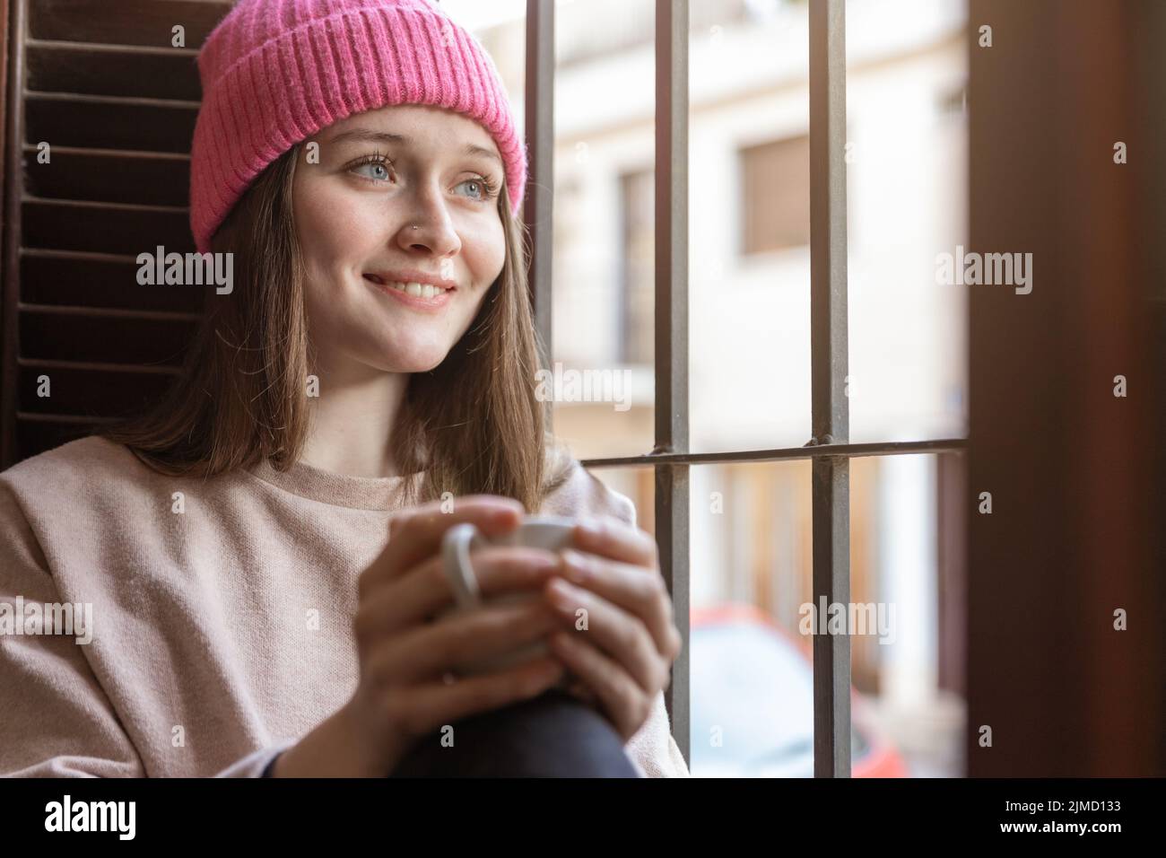 Jeune femme positive dans un chapeau avec une tasse de boisson chaude  donnant sur la fenêtre avec le visage rêveur tout en étant assise dans la  pièce lumineuse Photo Stock - Alamy
