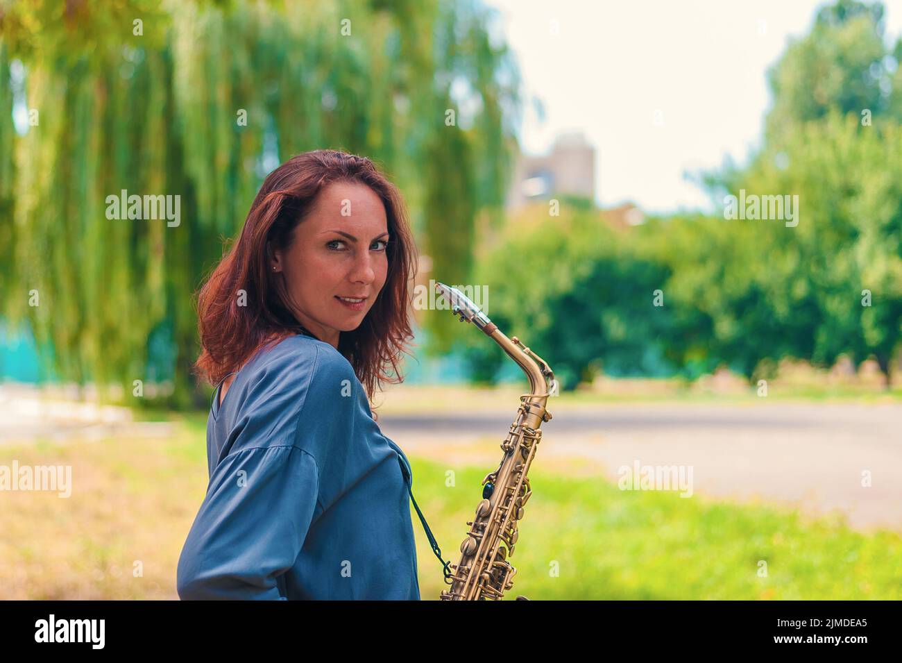 Jeune fille à tête rouge dans une veste bleue avec un saxophone souriant dans le parc Banque D'Images