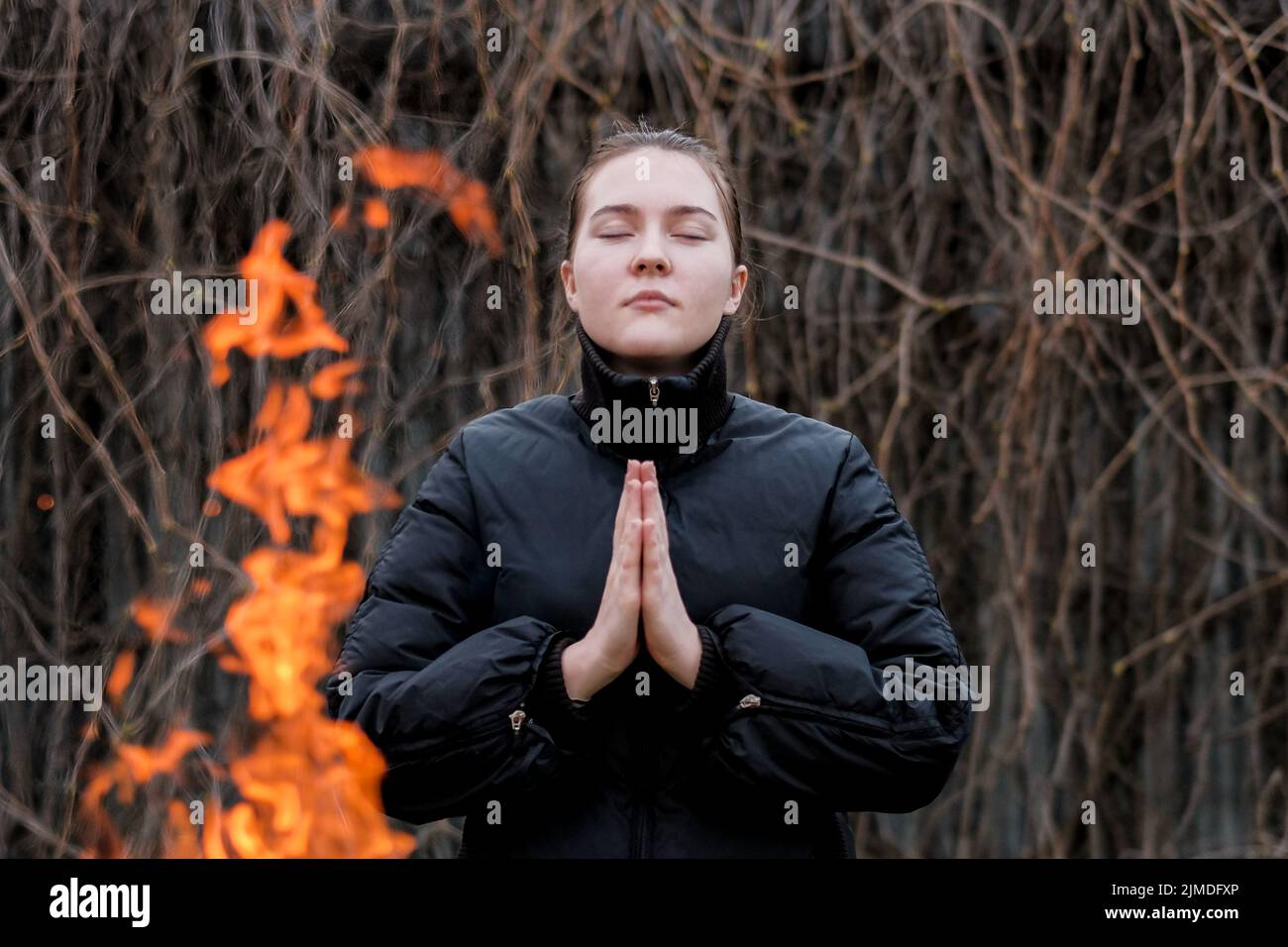 Une fille aux mains pliées dans la prière et aux yeux fermés est debout près du feu. Banque D'Images