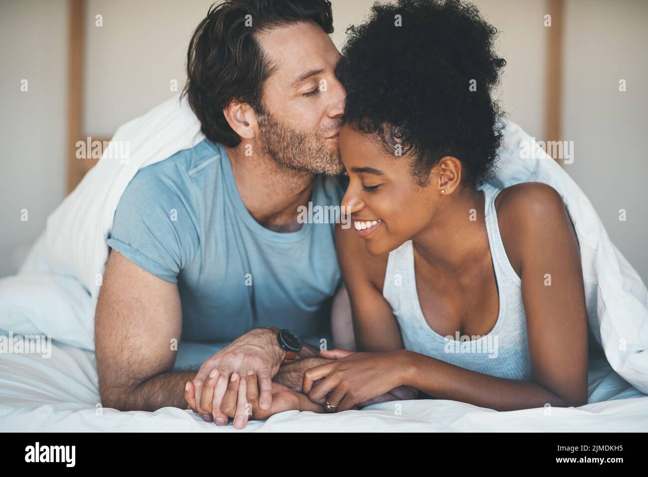 Un couple heureux, insouciant et aimant partage un baiser allongé dans le lit ensemble le matin. Liaison interraciale, intime et bienveillante de mari et de femme et Banque D'Images