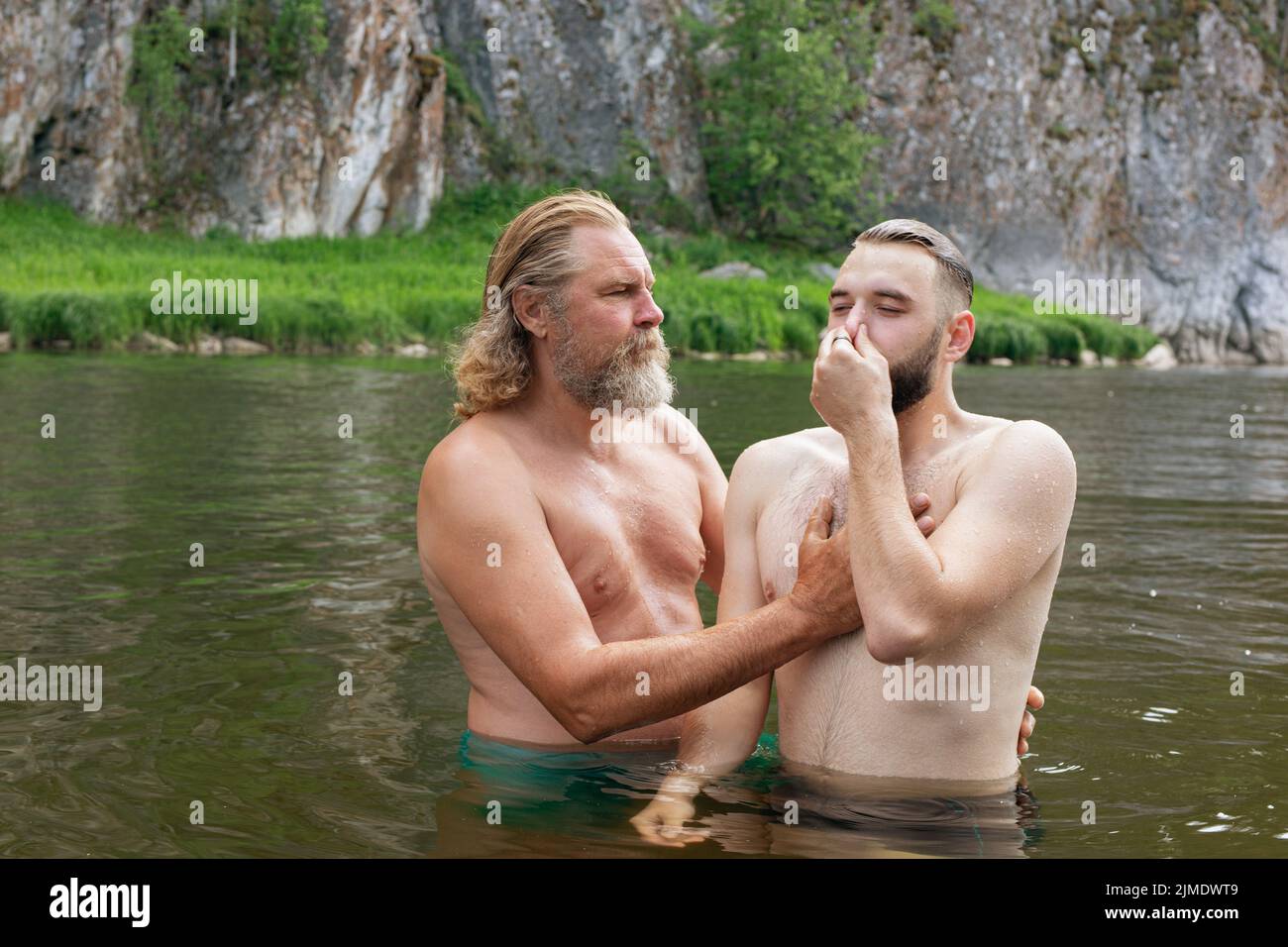 Deux hommes dans l'eau profonde à la taille, un jeune homme avec les yeux fermés et le nez pincé par la main écoute l'homme plus âgé. Cérémonie de baptême o Banque D'Images