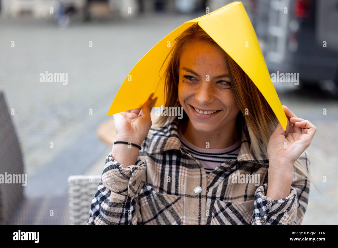 Une étudiante assise à l'extérieur sur une table de terrasse couvrant sa tête pour la pluie avec une chemise en plastique jaune Banque D'Images
