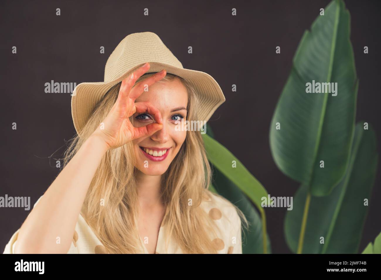 Femme blonde blanche dans un chapeau de soleil regardant à travers les doigts dans l'appareil photo avec le grand sourire. Geste parfait devant le visage. Grande plante. Prise de vue en studio à arrière-plan sombre. Photo de haute qualité Banque D'Images