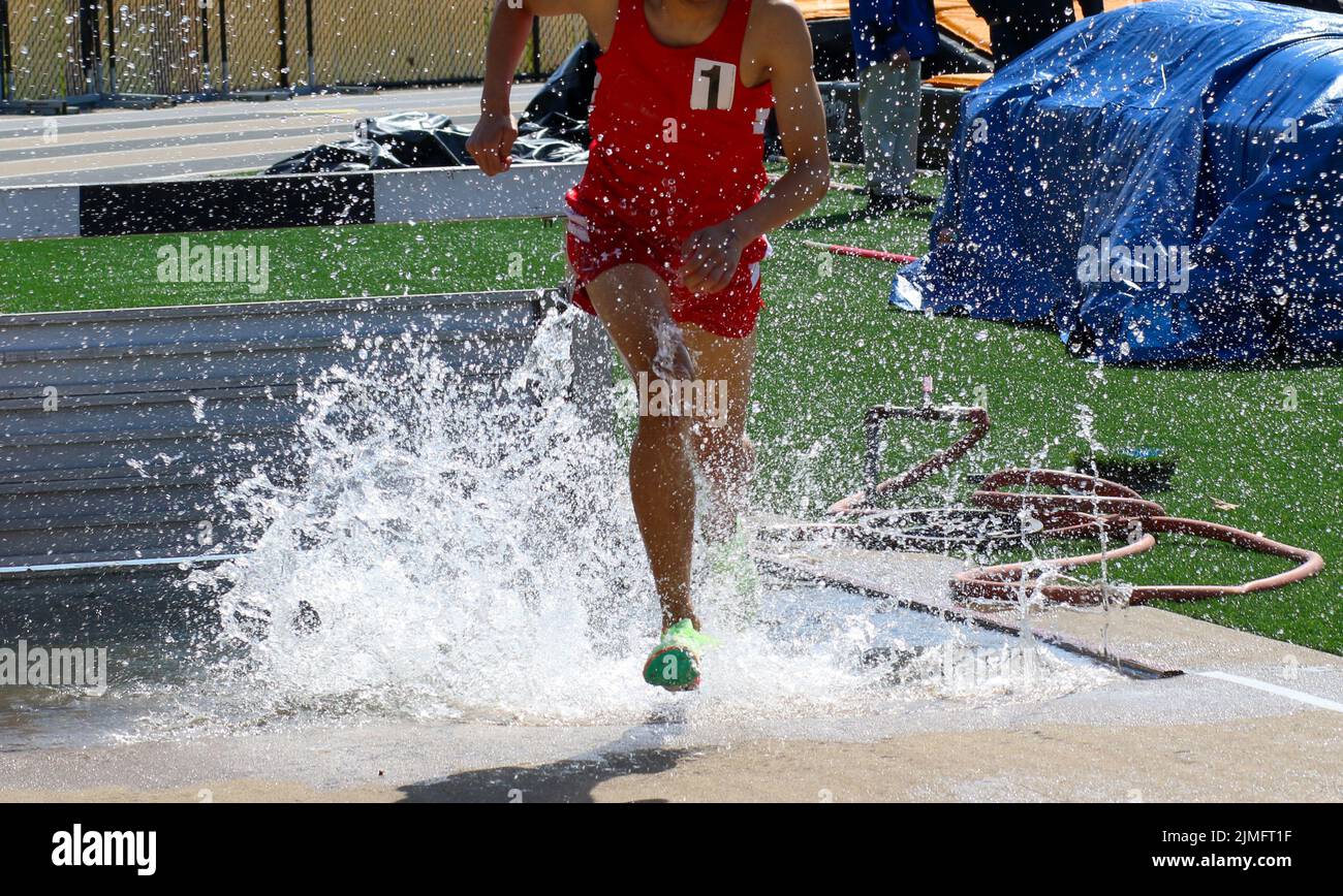 Un garçon du lycée éclaboussant lorsqu'il sort du saut à l'eau steeplechase pendant une course qu'il gagne. Banque D'Images