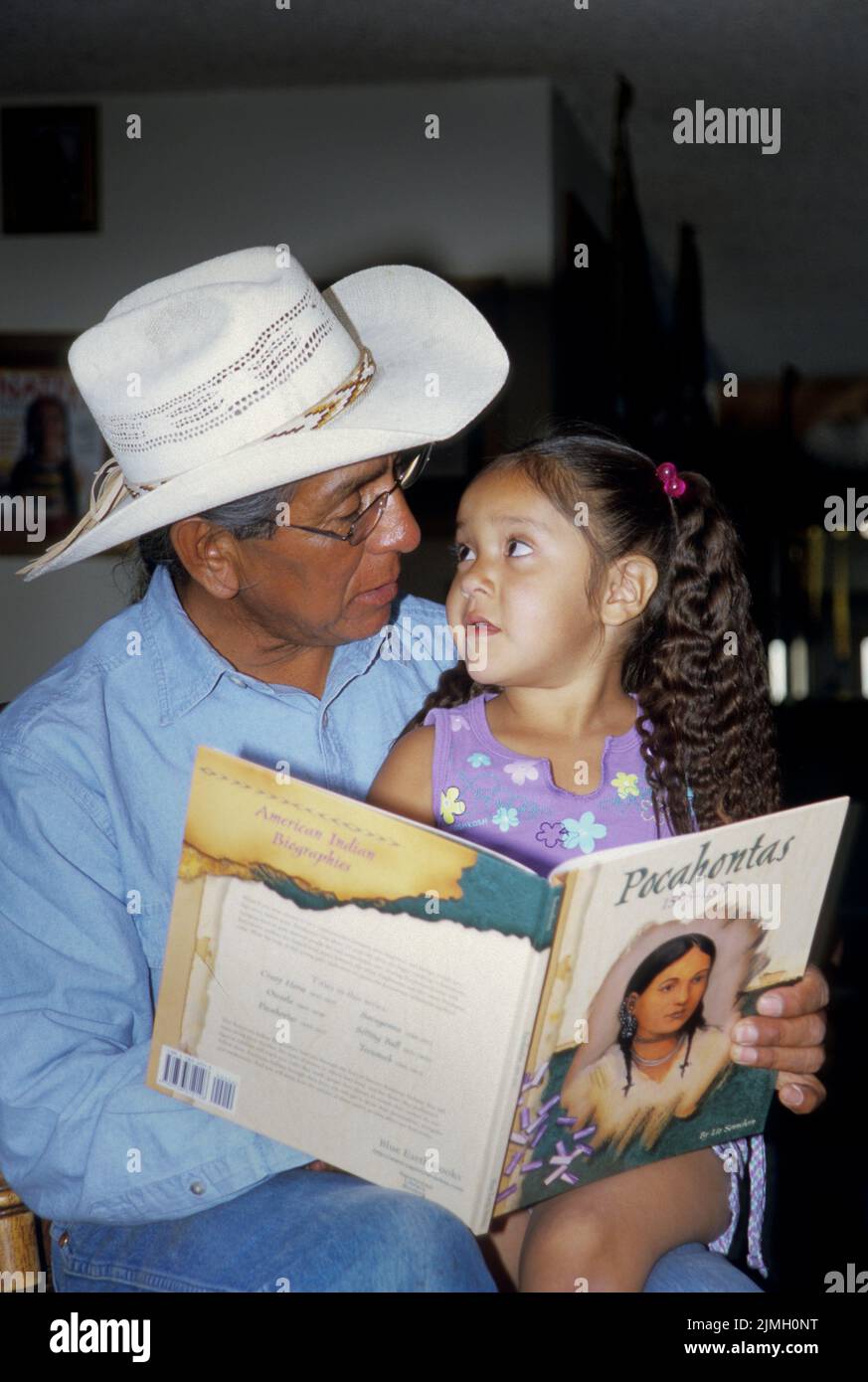 Famille amérindienne d'un grand-père lisant un livre d'histoire sur Pocahontas à sa petite-fille alors qu'elle le regarde. Banque D'Images