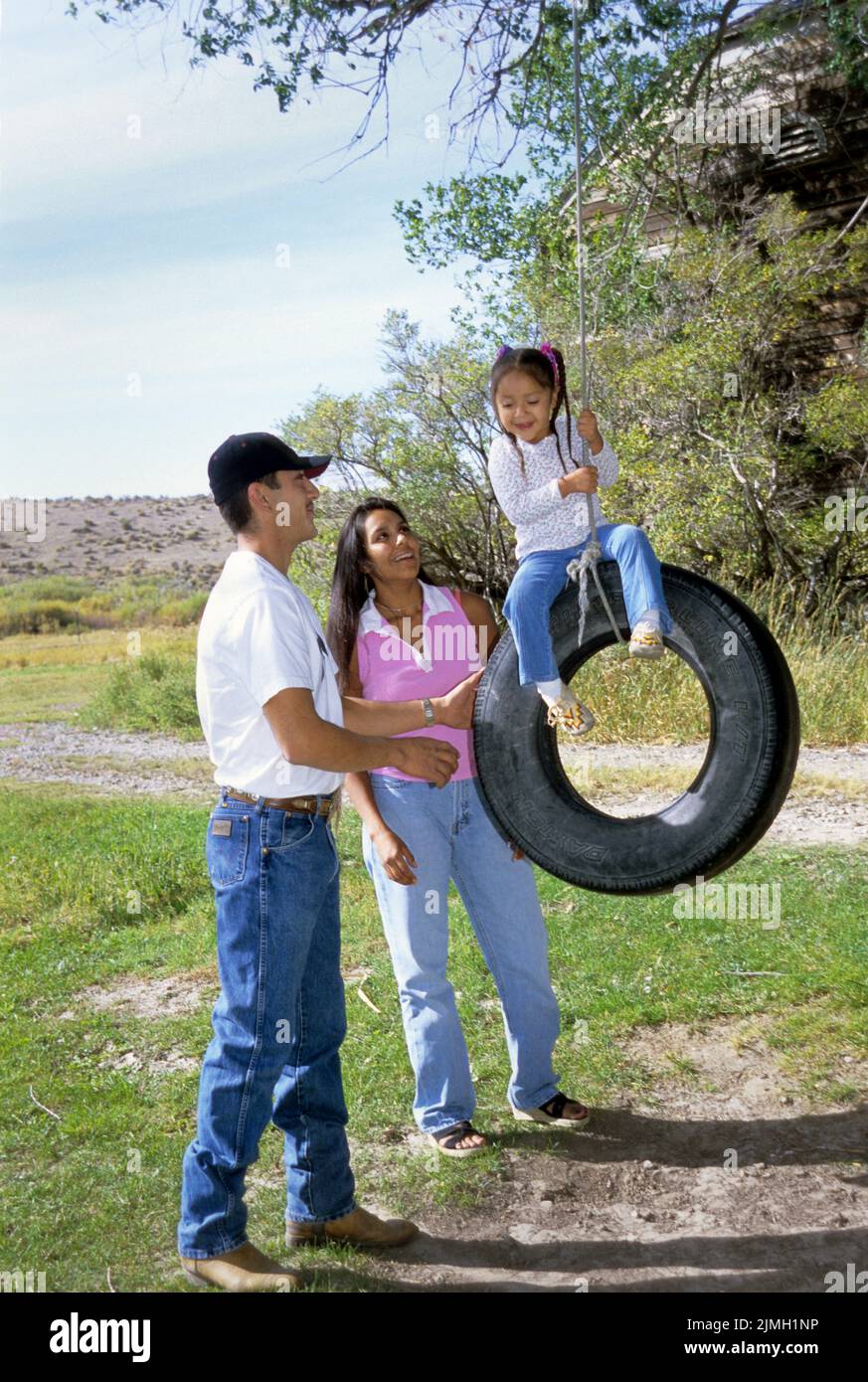 La famille native américaine de père et de mère avec une jeune fille joue avec une balançoire de pneu dans leur cour arrière Banque D'Images