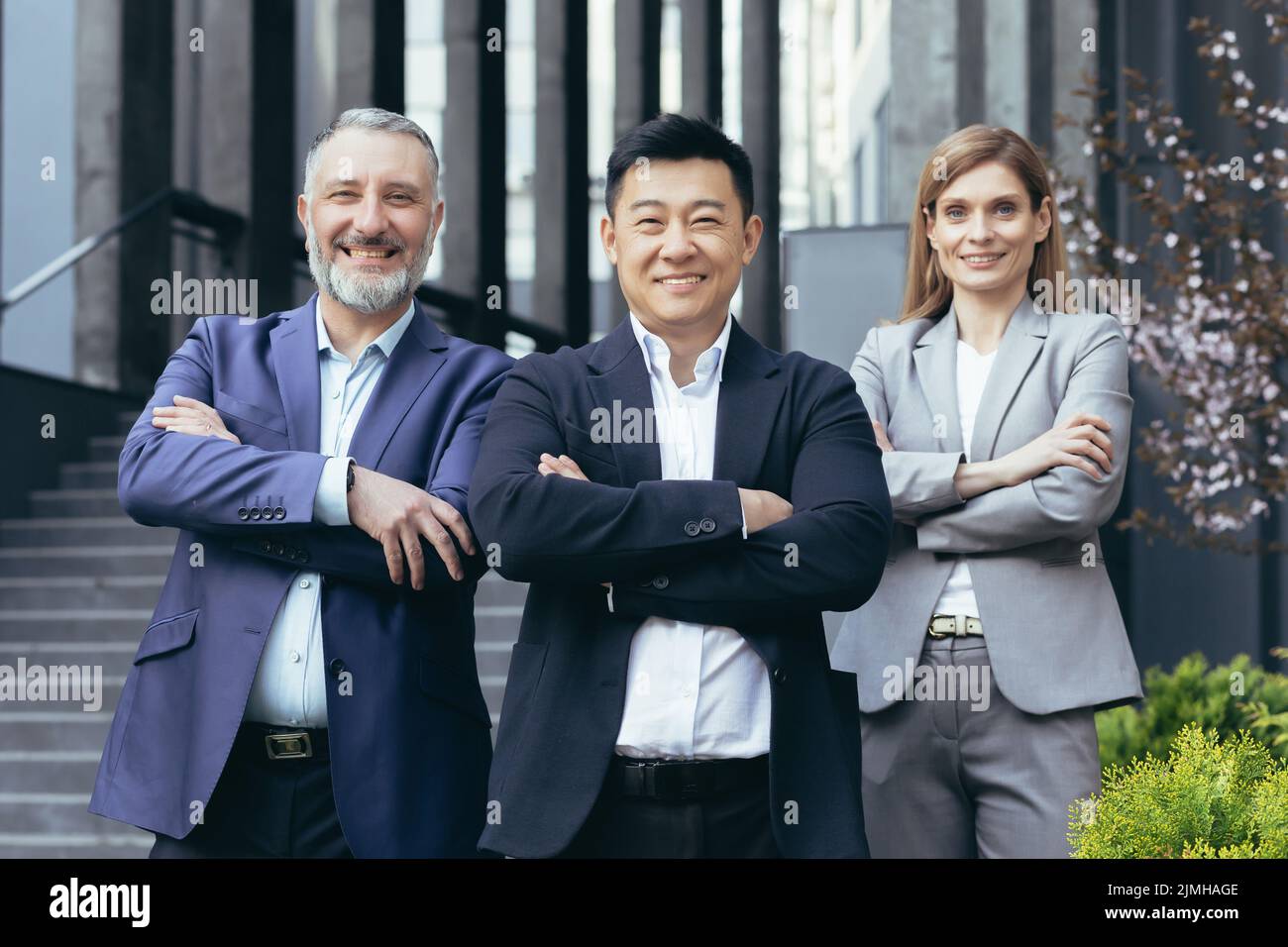 Portrait d'un patron asiatique réussi et heureux avec son équipe de rêve diversifiée, des gens d'affaires à l'extérieur du bâtiment de bureau debout avec les bras croisés et regardant l'appareil photo Banque D'Images