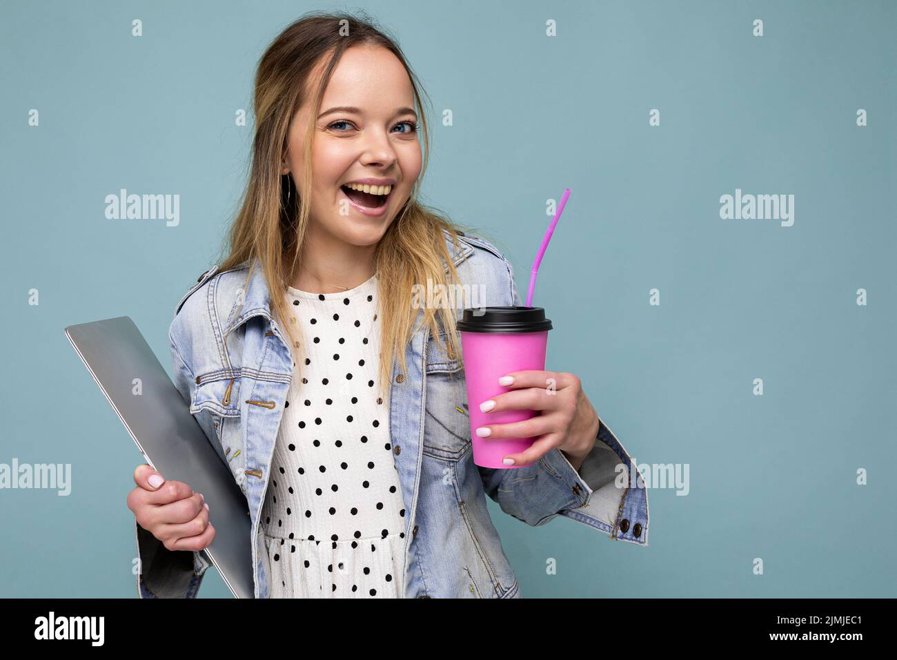 Image d'une joyeuse femme blonde européenne souriante et debout contre un mur bleu isolé d'un carnet argenté et d'un café à emporter Banque D'Images