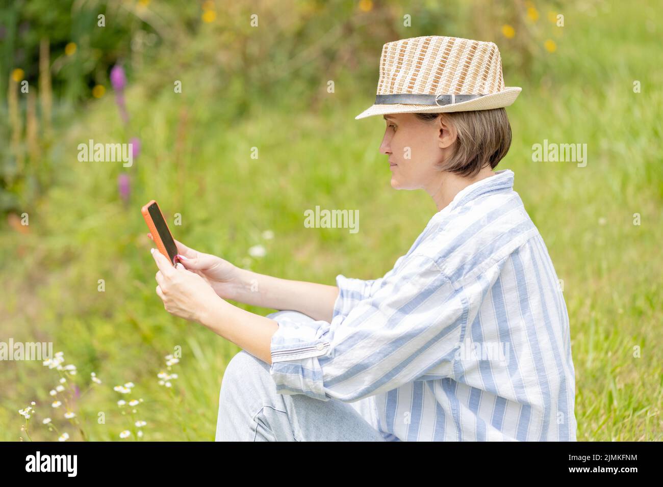 femme portant un chapeau assis dans le parc sur l'herbe verte avec un smartphone. travail à distance Banque D'Images
