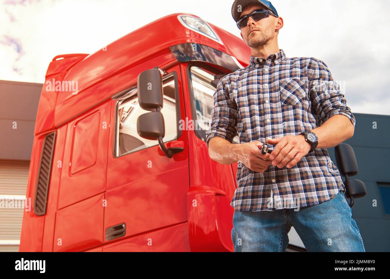 Caucasien Trucker dans son 40s fièrement debout devant la cabine rouge de son véhicule de marchandises lourdes attendant le chargement de partir. Transport et logistique Banque D'Images