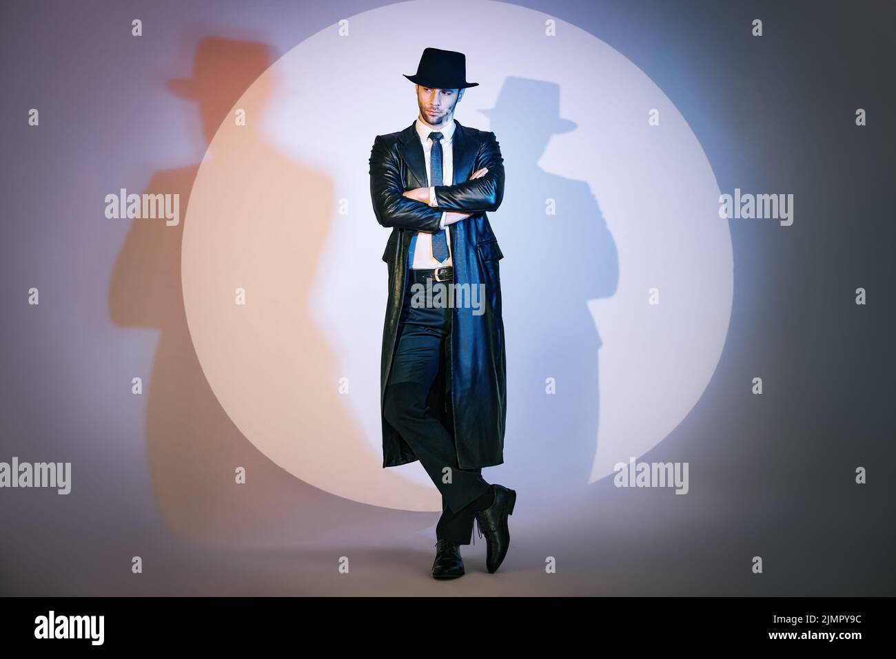 Portrait complet d'un homme doué et confiant en manteau noir et chapeau avec bras croisés posant sur le studio à la lumière du spot. Banque D'Images