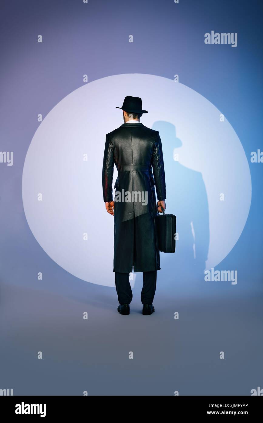 Vue arrière de la silhouette d'homme en manteau noir et chapeau tenant la mallette dans le projecteur sur l'arrière-plan du studio Banque D'Images