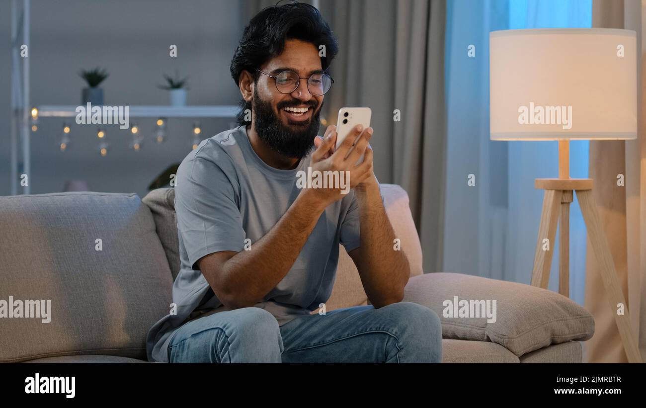 Joyeux homme millénaire indien arabe tenant un smartphone moderne avec un message texte à la maison assis sur un canapé le soir, un homme barbu dans des lunettes Banque D'Images