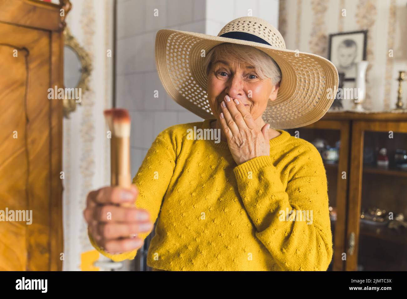 Drôle caucasien femme âgée, portant un chapeau de plage élégant à la maison et tenant des pinceaux de maquillage dans une main, l'aspect choqué et confus. Photo de haute qualité Banque D'Images