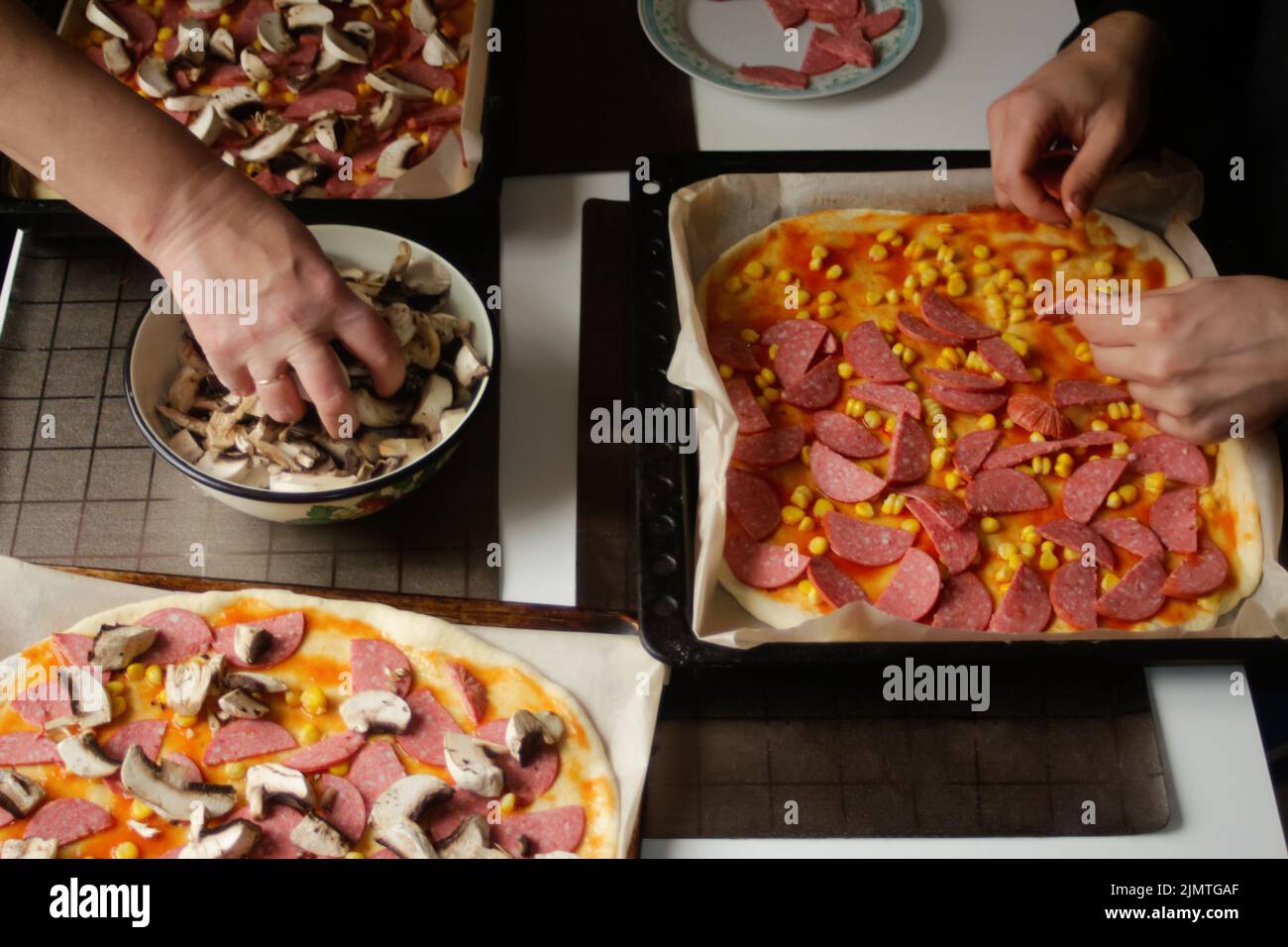 Permet de déconcentrer les ingrédients de la pizza sur la pâte. Femme et homme cuisine à la main. Sauce tomate rouge, saucisses, maïs doux, champignons. Pizza maison. Base de Marga Banque D'Images