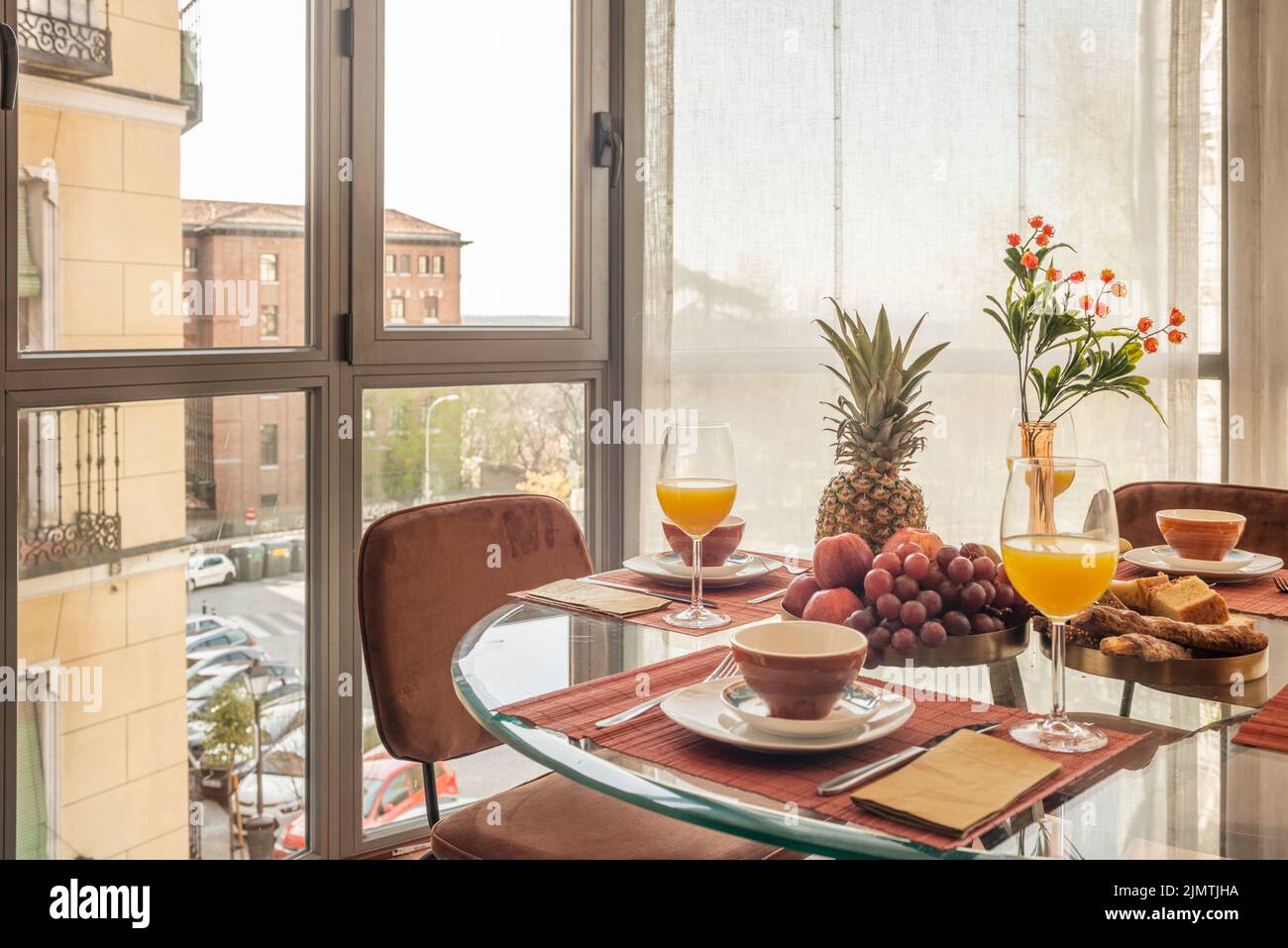 Salle à manger avec une table circulaire en verre pleine de fruits variés, des verres de jus d'orange et des chaises brunes à côté d'une baie vitrée avec vue Banque D'Images