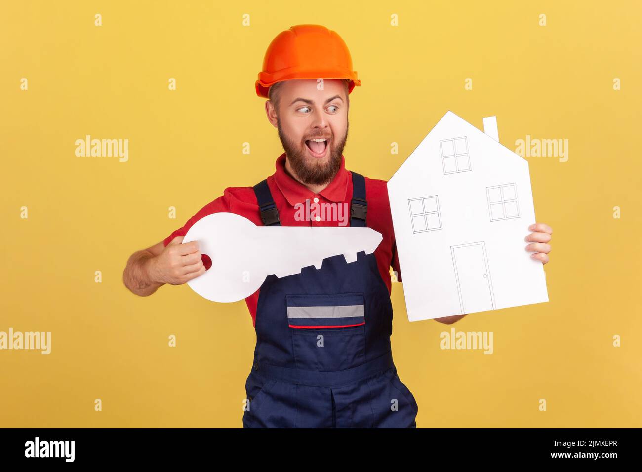 Portrait d'un travailleur enthousiaste tenant une grosse clé en papier et une maison entre les mains, hurler quelque chose d'excitante, porter une combinaison et une casquette rouge. Studio d'intérieur isolé sur fond jaune. Banque D'Images