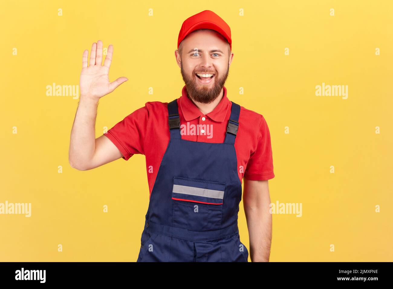 Portrait d'un ouvrier ravi debout et regardant l'appareil photo, en agitant la main, en disant bonjour ou Au revoir, en portant une combinaison bleue et une casquette rouge. Studio d'intérieur isolé sur fond jaune. Banque D'Images