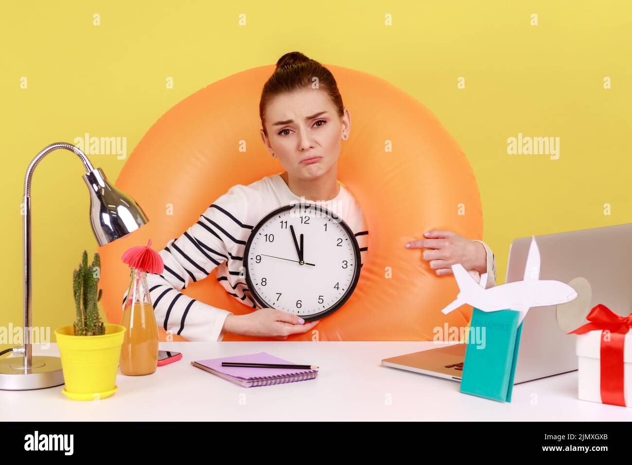 Triste femme bouleversée dans l'anneau en caoutchouc sur le cou assis sur le lieu de travail avec un ordinateur portable, montrant l'horloge murale, n'a pas le temps d'aller en vacances d'été. Studio d'intérieur tourné isolé sur fond jaune. Banque D'Images