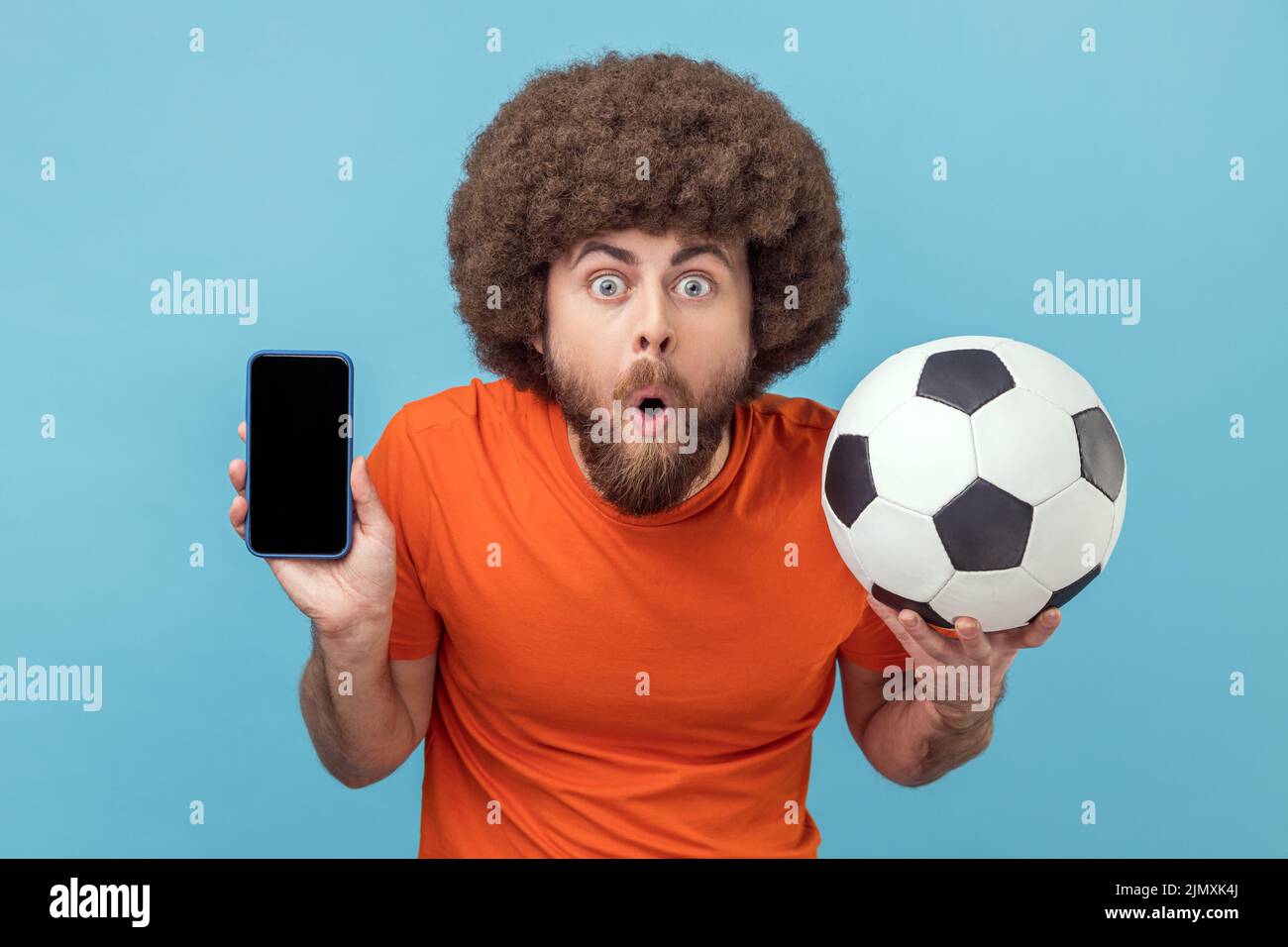 Portrait d'un homme choqué avec une coiffure afro portant un T-shirt debout avec une balle de football et montrant un téléphone portable avec un affichage vide pour la publicité. Studio d'intérieur isolé sur fond bleu. Banque D'Images
