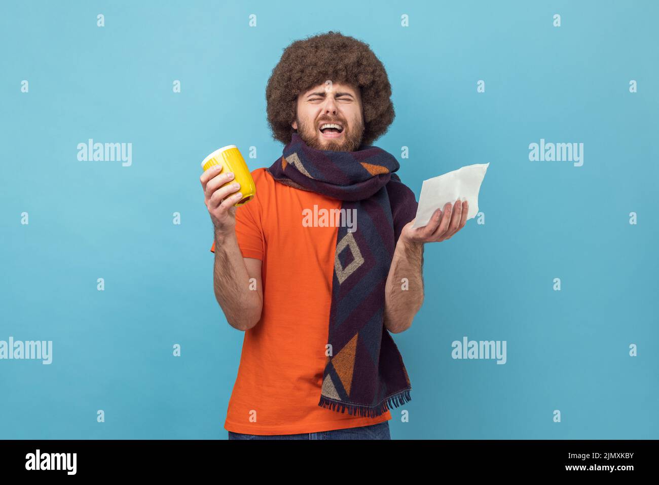 Portrait d'un homme avec une coiffure afro en T-shirt orange tenant une tasse et un tissu pour l'écoulement nasal, prenant des médicaments avec le thé chaud, la fièvre de souffrance, les maux de tête. Studio d'intérieur isolé sur fond bleu. Banque D'Images