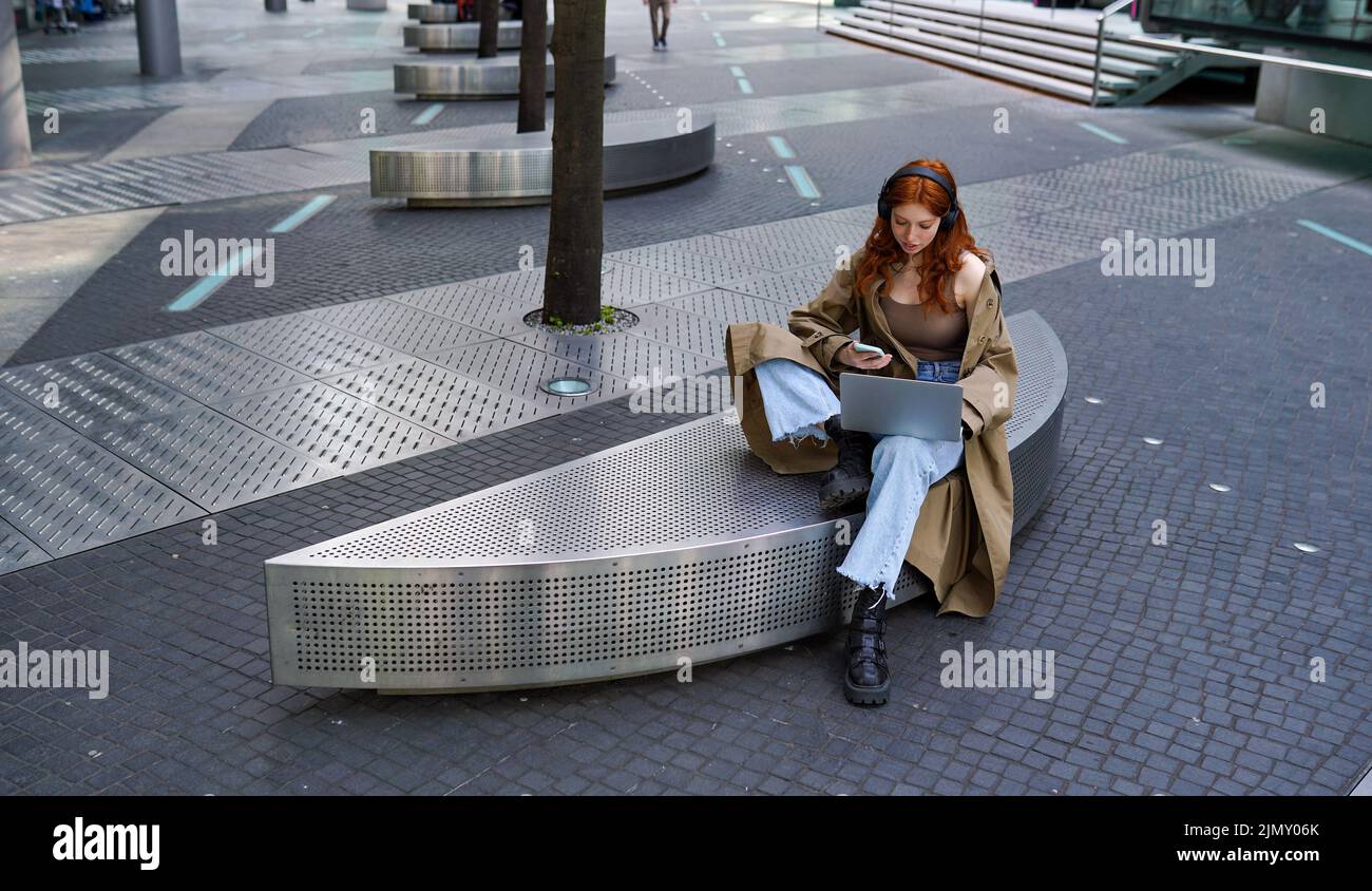 Ado redhead hipster fille utilisant un ordinateur portable et téléphone dans la rue urbaine de la ville. Banque D'Images